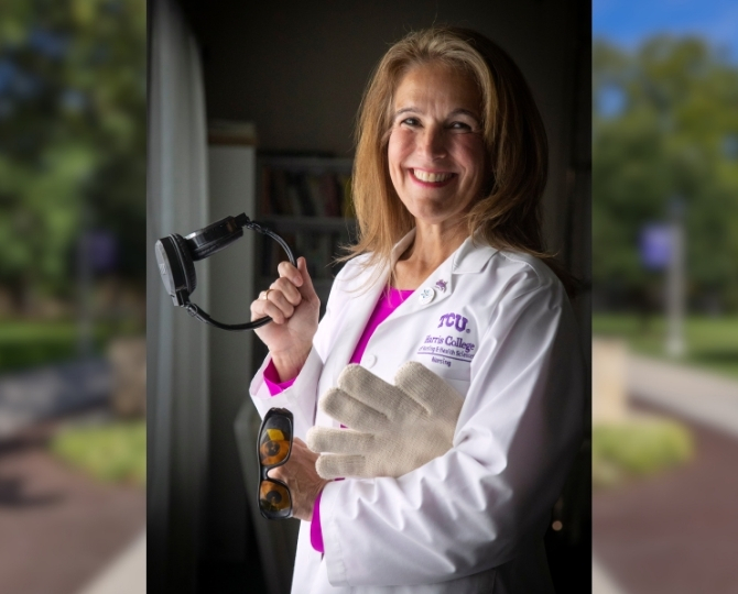 A smiling woman wearing a white TCU Harris College of Nursing & Health Sciences lab coat stands indoors near a window, holding a handheld examination light in one hand and a pair of protective glasses in the other. She is also wearing a lead protective glove and looks directly at the camera. The softly blurred background keeps the focus on her professional attire and clinical equipment.