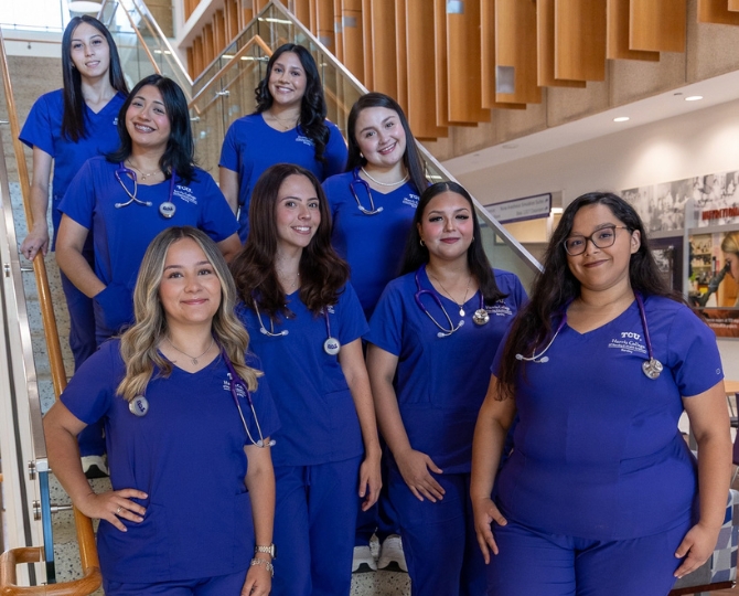 8 nursing students in purple scrubs stand on a staircase, smiling, wearing stethoscopes around their necks.